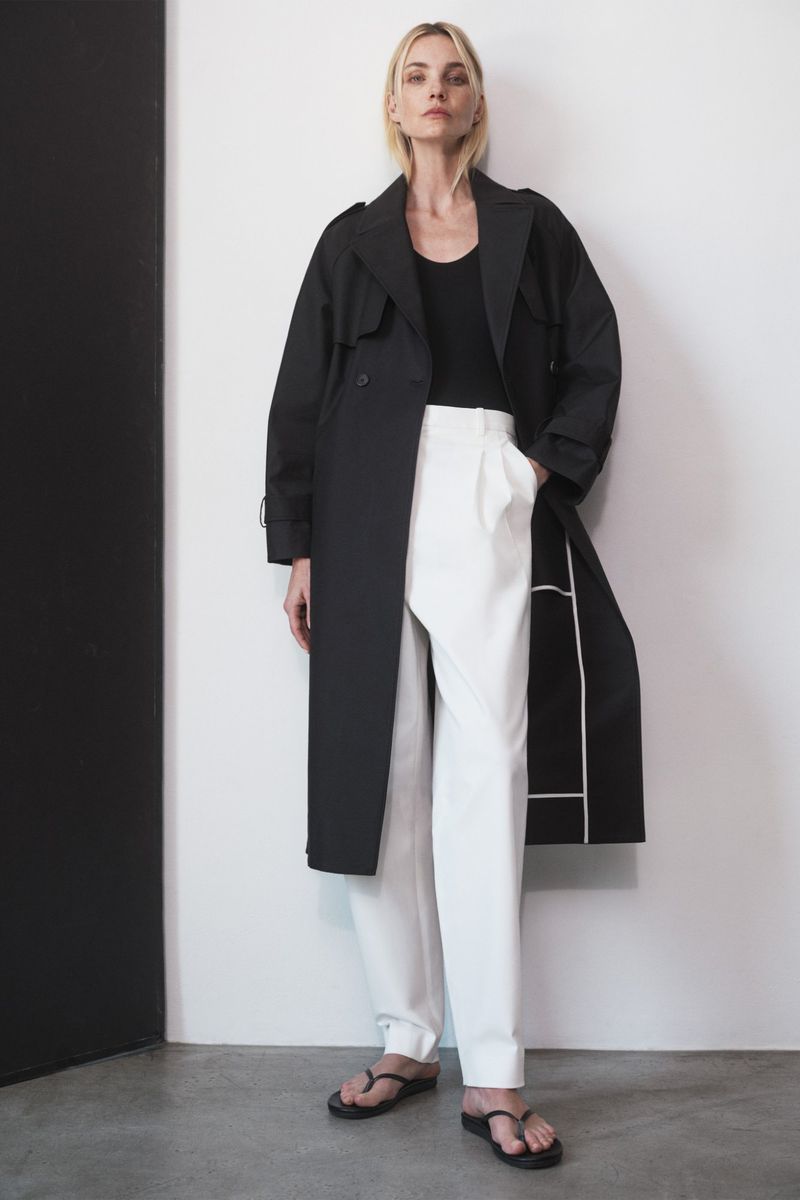 Full-shot of a stylish woman in a black trench coat and white trousers, posing indoors with neutral expression and minimal accessories.