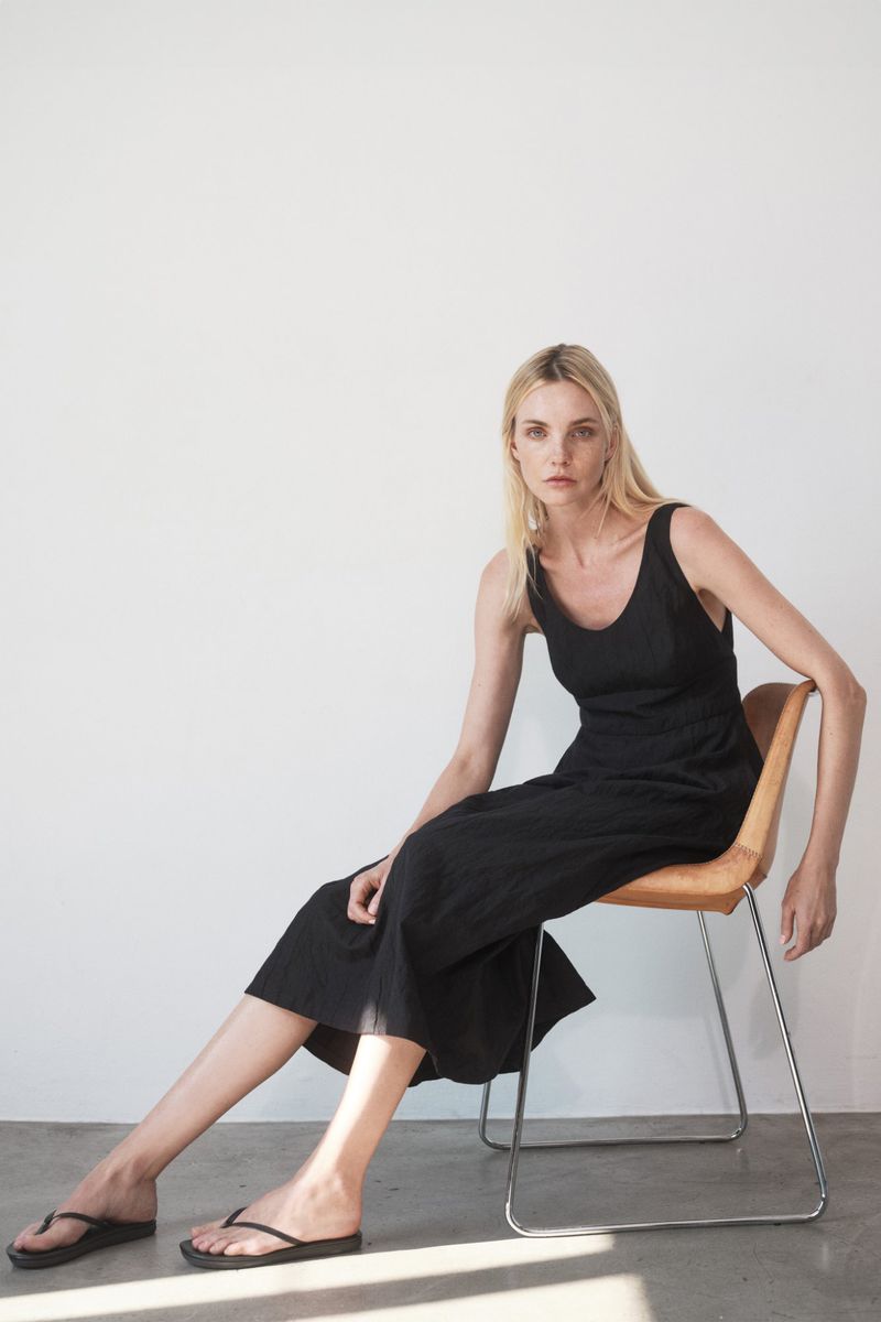 An adult woman sits on a chair in a photo studio, wearing a black dress and flip-flops against a bright white background, indoors.