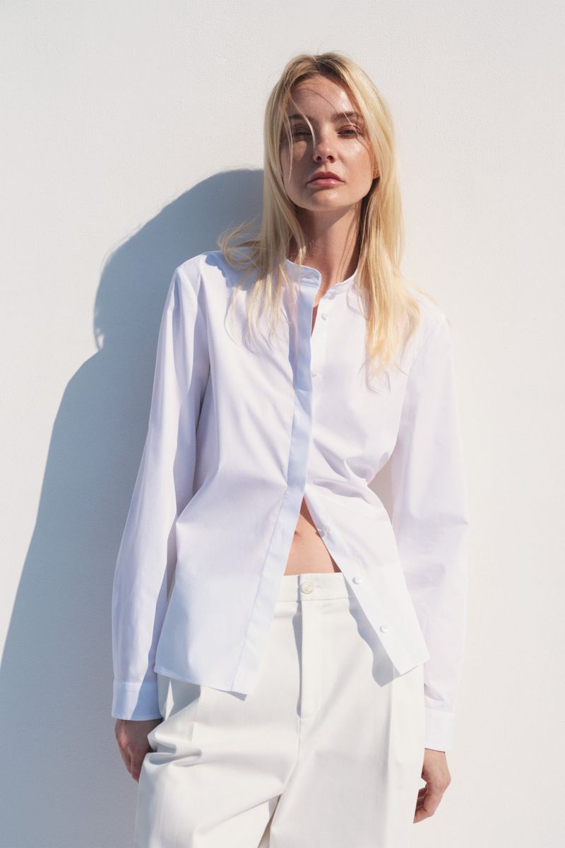 A fair-skinned woman with blonde hair poses against a white wall in an unbuttoned white shirt and pants in a fashion shot.