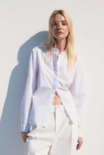 A fair-skinned woman with blonde hair poses against a white wall in an unbuttoned white shirt and pants in a fashion shot.