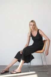 Model Caroline Trentini seated on a tan modern chair wearing a black sleeveless midi dress and flip-flops against a white wall.