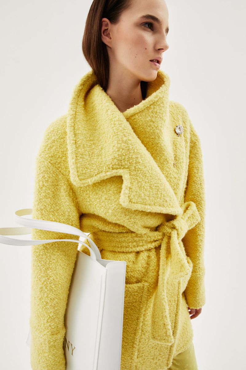 A fair-skinned young woman poses in a yellow belted jacket, holding a white shopping bag in a studio with a clean, bright background.