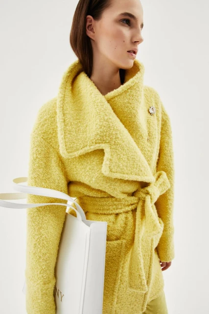 A fair-skinned young woman poses in a yellow belted jacket, holding a white shopping bag in a studio with a clean, bright background.