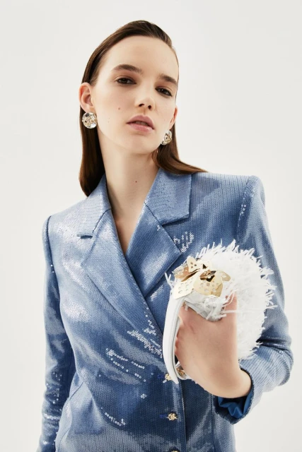 Woman in a blue sequined blazer poses against a white backdrop, holding a white feathered handbag in a medium, eye-level shot.