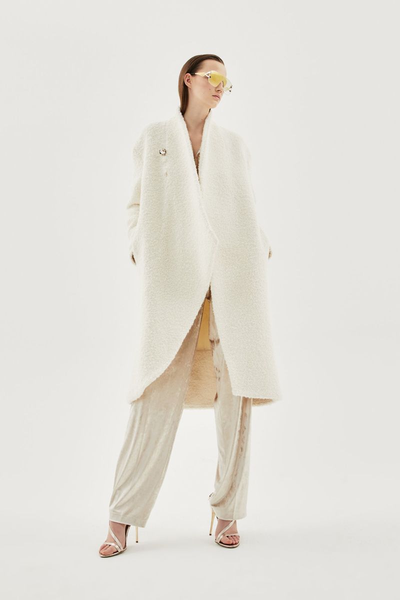 Model Emma Muszela wearing a long white textured coat and cream trousers with yellow shield sunglasses on a white studio background.