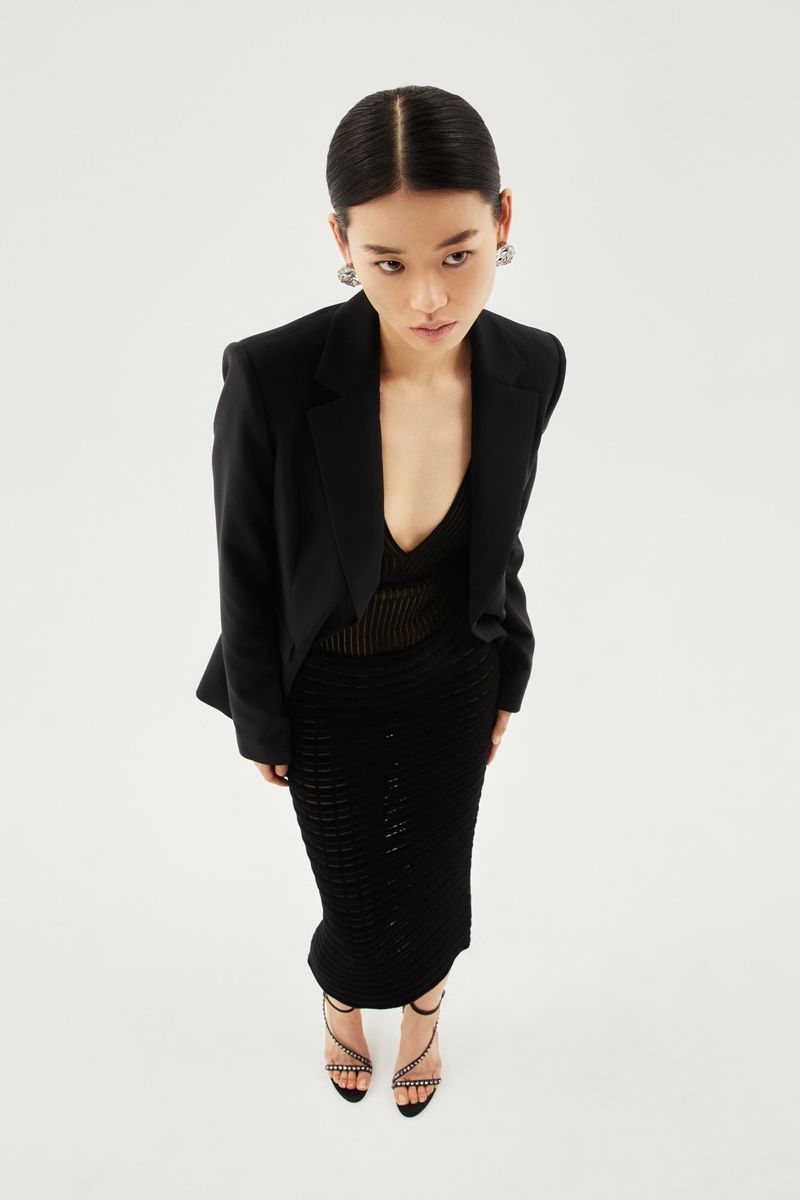 High-angle view of model Emma Muszela in a black blazer, black midi skirt, and strappy heels against a white background.