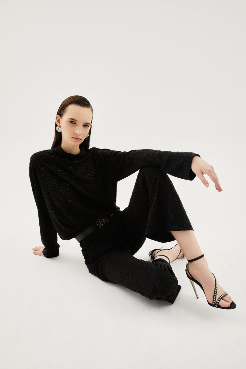 Elegant adult woman in a black sweater and pants sitting on a white backdrop, wearing high-heeled sandals and a black belt. Fashion shot.