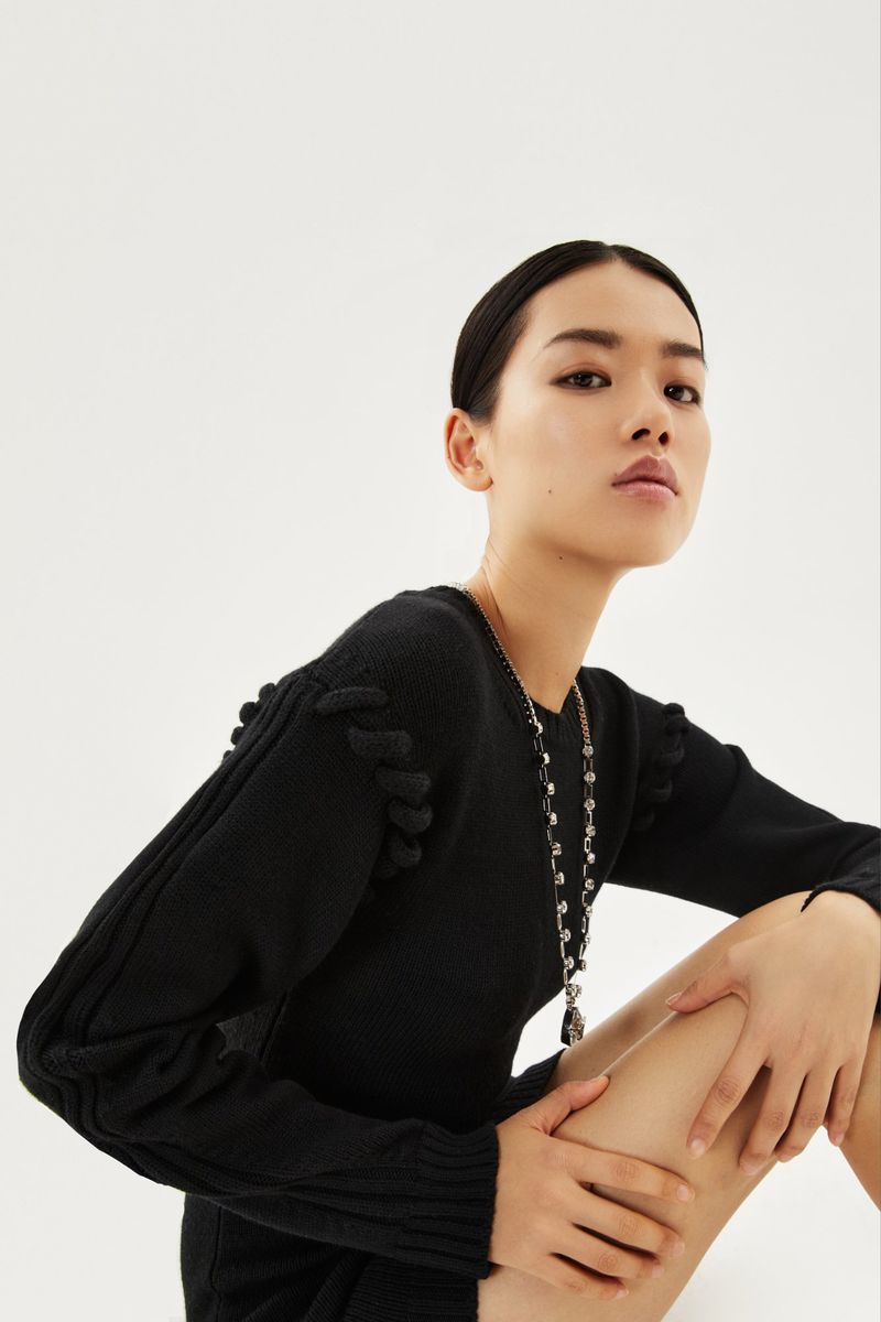 A young Asian female fashion model poses in a black knit dress against a clean white backdrop, showcasing modern fashion and style.