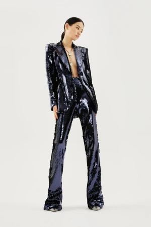 Model Emma Muszela wearing a glittering navy blue sequined pantsuit and necklace against a plain off-white studio background.