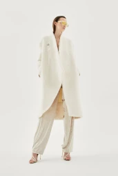 Model Emma Muszela wearing a long white textured coat and cream trousers with yellow shield sunglasses on a white studio background.