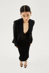High-angle view of model Emma Muszela in a black blazer, black midi skirt, and strappy heels against a white background.