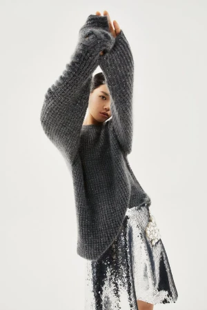 Model Emma Muszela wearing an oversized grey knit sweater and silver sequin skirt against a white studio background.