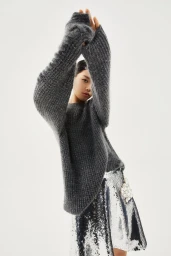 Model Emma Muszela wearing an oversized grey knit sweater and silver sequin skirt against a white studio background.