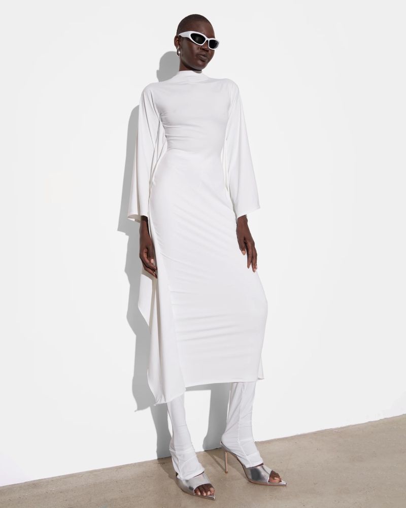 A slender dark-skinned woman poses in a long-sleeved white dress, matching leggings, silver heels and white sunglasses against a white background.