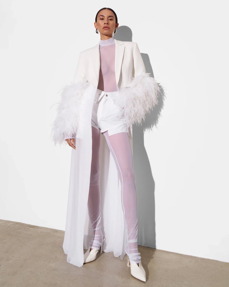 A stylish adult female poses in a white blazer with feather sleeves, white shorts, and sheer leggings against a neutral backdrop.
