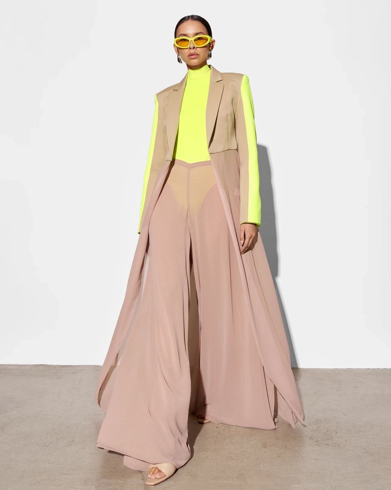 Fashionable woman in beige trench coat and wide-leg pants with neon yellow top and glasses stands confidently against a white backdrop.