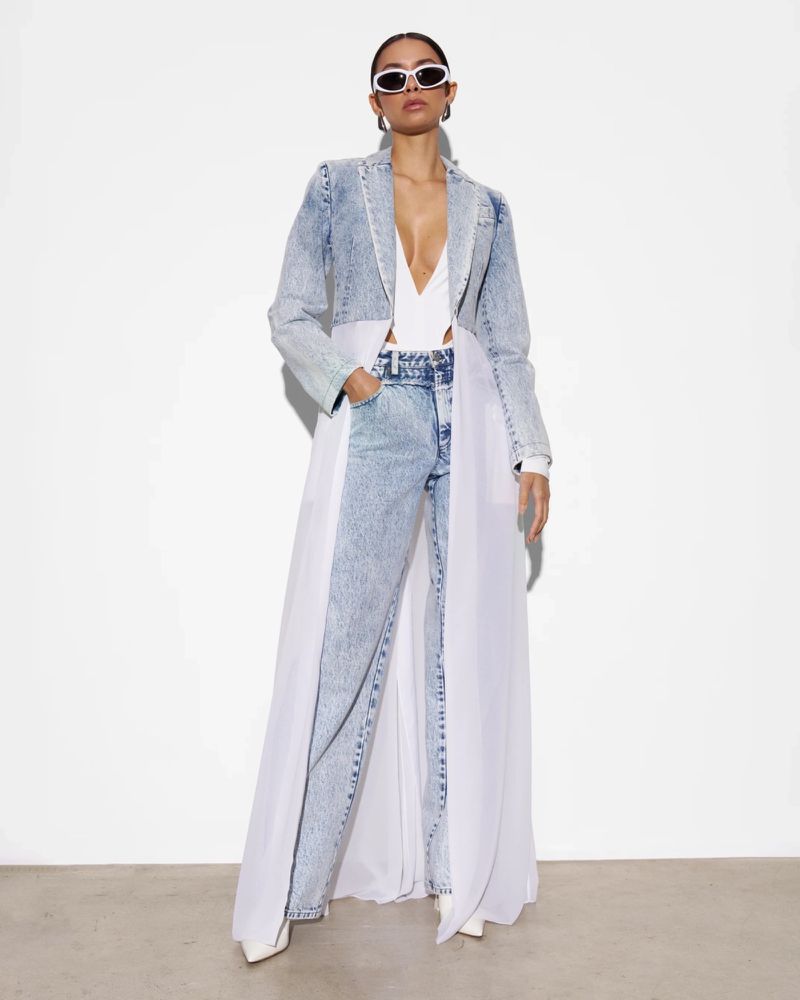 A woman poses in a denim coat and jeans with white sheer accents, complemented by white sunglasses and pointed heels against a white backdrop.