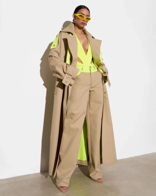 Woman poses in long beige trench coat, neon-yellow top, and pants against a white backdrop, accessorized with yellow glasses and earrings.