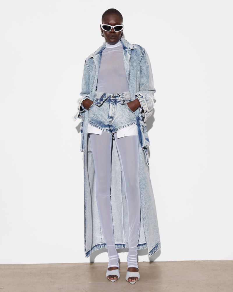 Editorial fashion shot of a woman with dark skin in a denim coat, shorts, and sheer leggings, wearing sunglasses against a white backdrop.