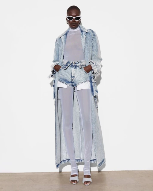 Editorial fashion shot of a woman with dark skin in a denim coat, shorts, and sheer leggings, wearing sunglasses against a white backdrop.