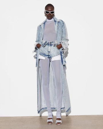 Editorial fashion shot of a woman with dark skin in a denim coat, shorts, and sheer leggings, wearing sunglasses against a white backdrop.