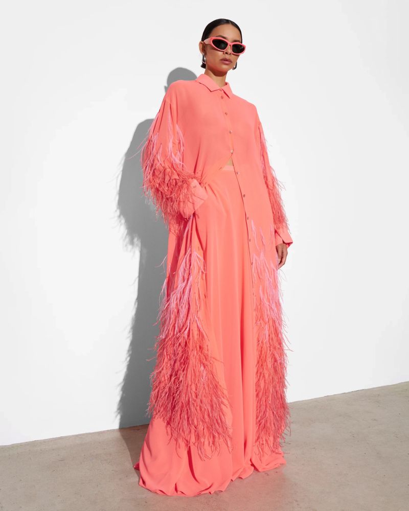A fashion model poses in a long, salmon-colored, feather-trimmed dress and retro sunglasses against a minimalist off-white backdrop.