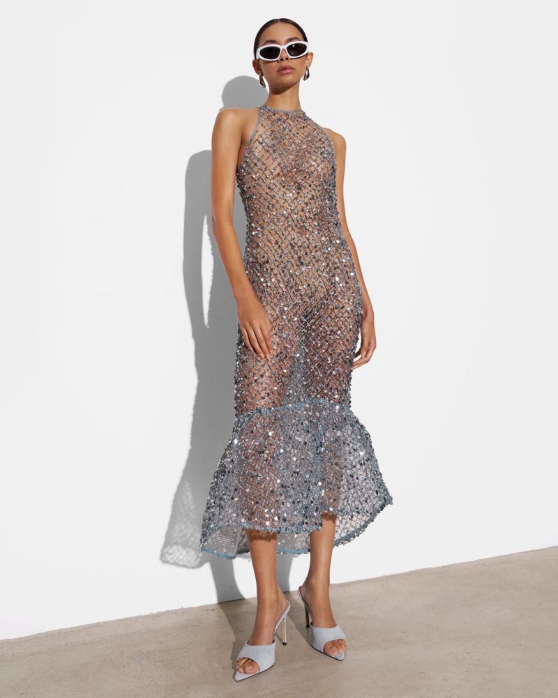 Elegant adult female fashion model poses in a sheer, embellished halter dress and sunglasses against a white wall in a photo studio.
