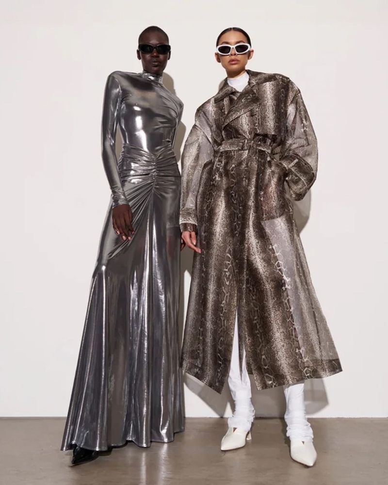 Two women pose in a studio wearing fashionable attire. One wears a metallic silver dress, and the other a snakeskin trench coat.