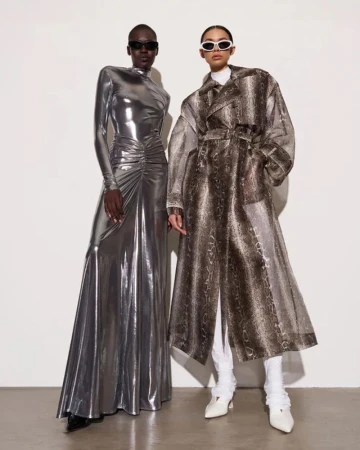 Two women pose in a studio wearing fashionable attire. One wears a metallic silver dress, and the other a snakeskin trench coat.