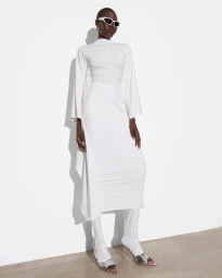 Model Nanny Malual in a draped white long-sleeve dress, matching leggings, futuristic sunglasses, and silver heels against a white wall.