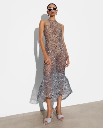 Model in white oval sunglasses wearing a sheer silver sequin halter midi dress and light blue mule heels against a white wall.