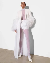 Model wearing white feathered coat, sheer lilac top, white shorts, and white heeled shoes against plain wall.