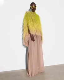 Model wearing a textured lime-yellow feathered top and flowing blush trousers with yellow sunglasses against a white wall.