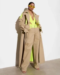 Model wearing an oversized tan trench coat, wide-leg trousers, neon lime top, and yellow sunglasses against a white wall.