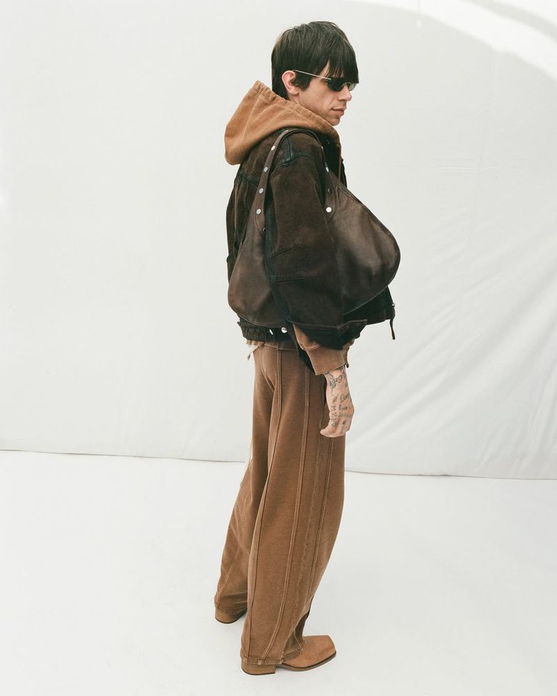 Model Cole Mohr standing in profile wearing a brown hoodie, wide-leg trousers, and carrying an oversized shoulder bag against a white backdrop.
