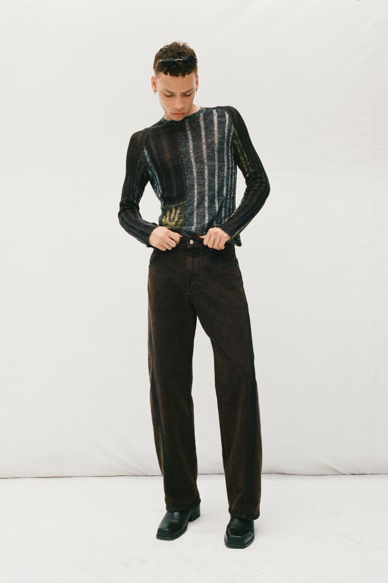 Studio shot of a young adult male model wearing a multi-colored knit sweater, dark corduroy pants, and black leather shoes, looking down.