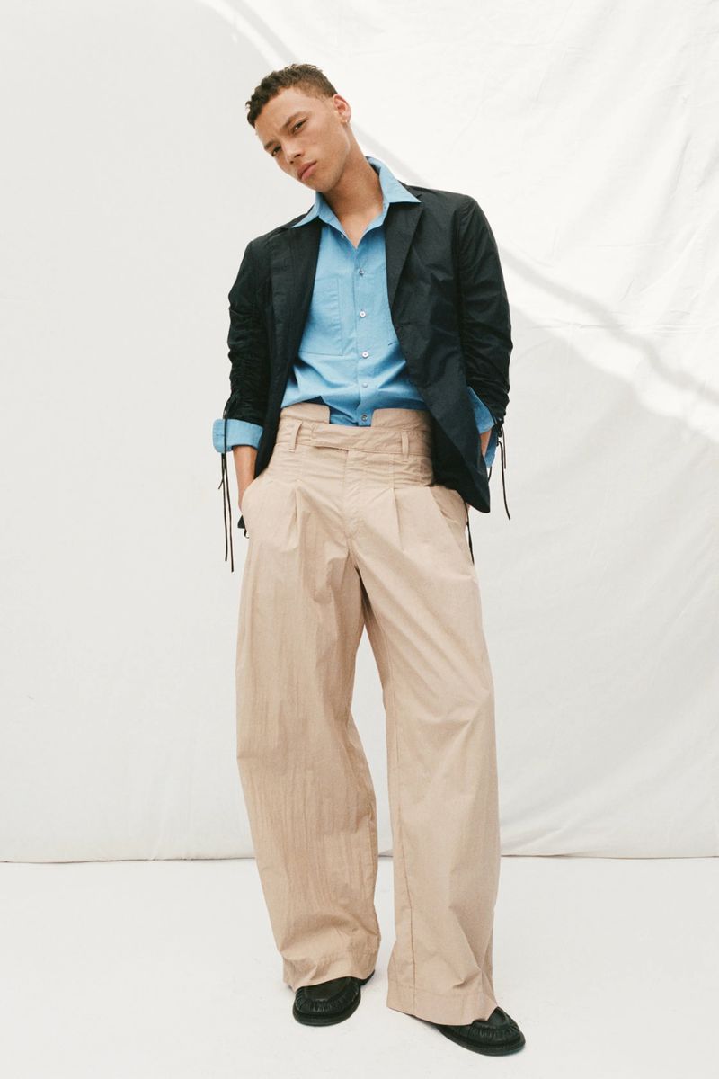 Male fashion model posing in a blue dress shirt, dark blazer, and oversized tan pants against a white backdrop in a fashion editorial.