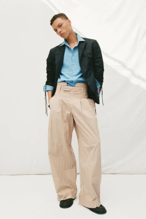 Male fashion model posing in a blue dress shirt, dark blazer, and oversized tan pants against a white backdrop in a fashion editorial.