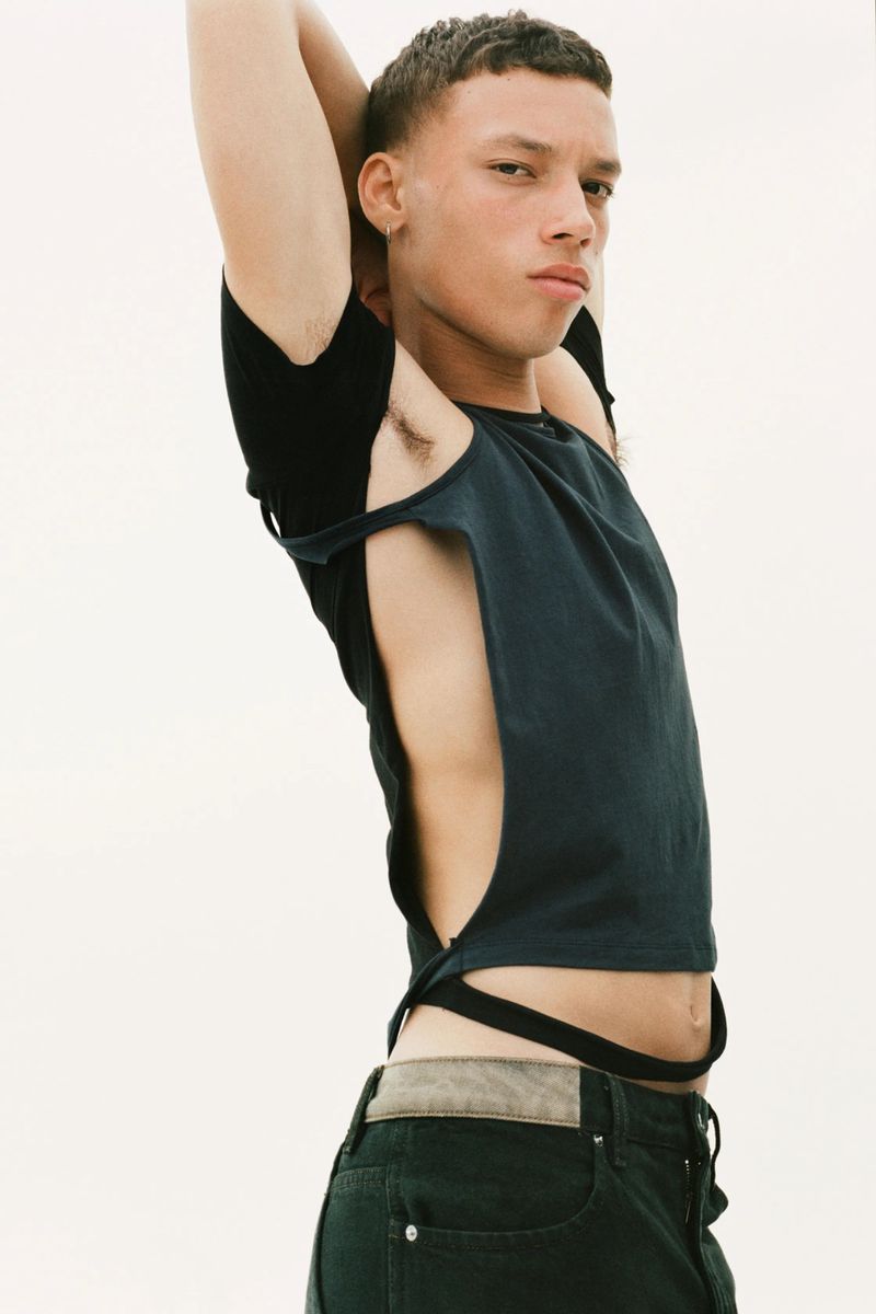 Portrait of a male model wearing a cut-out black shirt and dark jeans. The model has short hair and arms raised, against a neutral background.
