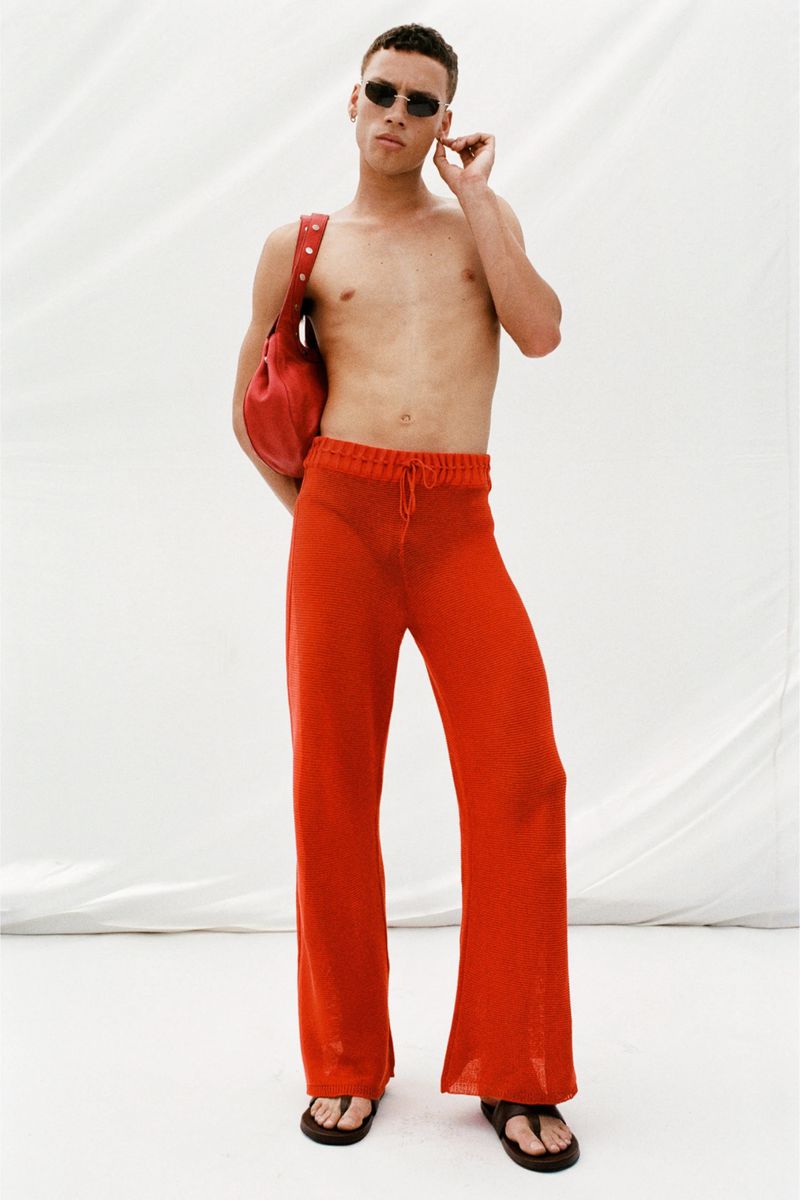 A model stands in a white studio, wearing a red see-through knit outfit and sunglasses, with a red leather bag slung over the shoulder.