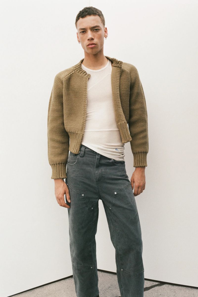 A young adult male stands in a studio wearing a tan knit jacket, white t-shirt, and dark grey denim pants looking at the viewer.