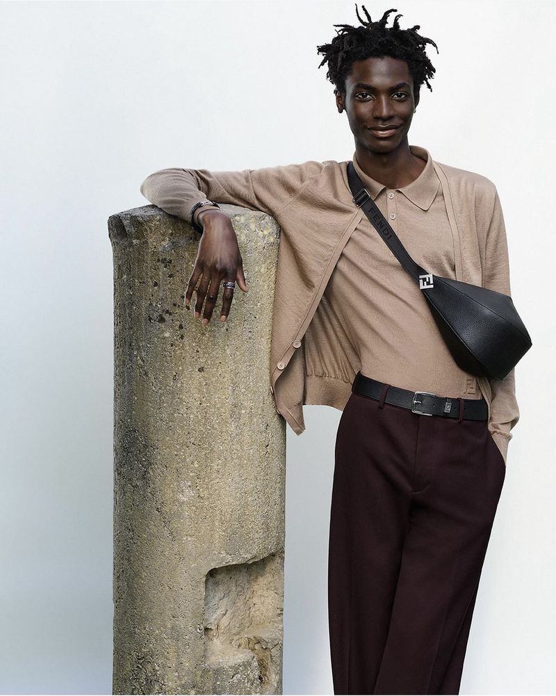 An adult male model poses in earth-tone fashion with a black bag and brown trousers, leaning against a grey textured column.