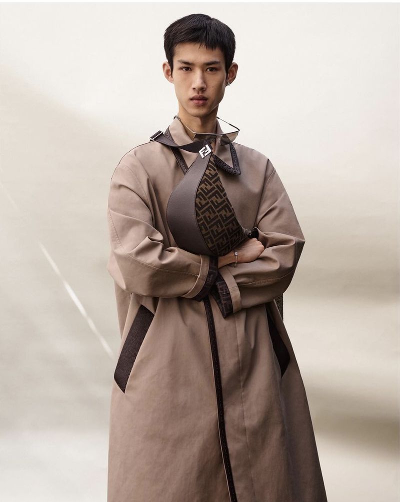 Asian male model posing with a brown patterned Fendi bag and beige trench coat against a neutral backdrop in a studio setting.
