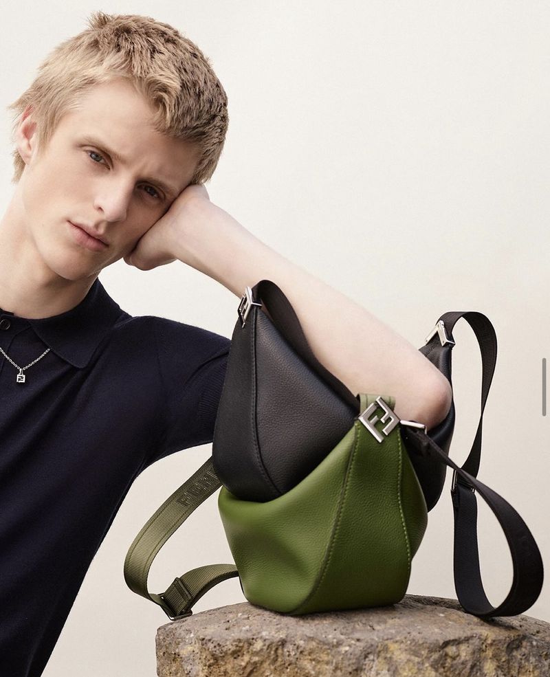 Fashion shot of a blond male model posing with a black and green purse placed on a stone surface against a plain background.