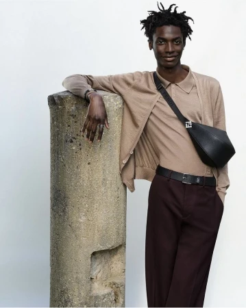 Model leaning against a stone pillar wearing a beige knit cardigan, matching polo, dark trousers, and a black crossbody bag.