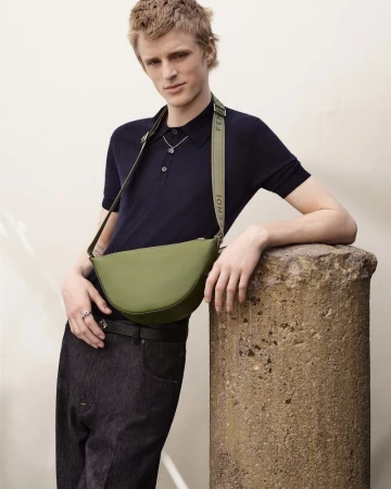 Model wearing a navy polo shirt and dark trousers leaning on a stone column with a green leather crossbody bag for Fendi.