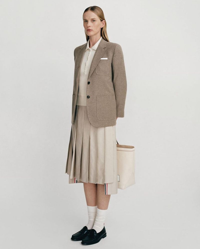 A tall, slender woman poses in a beige wool blazer, cream-colored pleated skirt, white socks, and black loafers against a neutral backdrop.