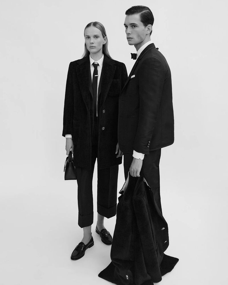 Two fashion models, a male and a female, pose in a black and white studio shot, showcasing formal wear, including a blazer, dress shoes and handbag.