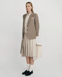 Model wearing a tailored brown blazer, pleated skirt, and loafers with a handbag against a seamless white background.