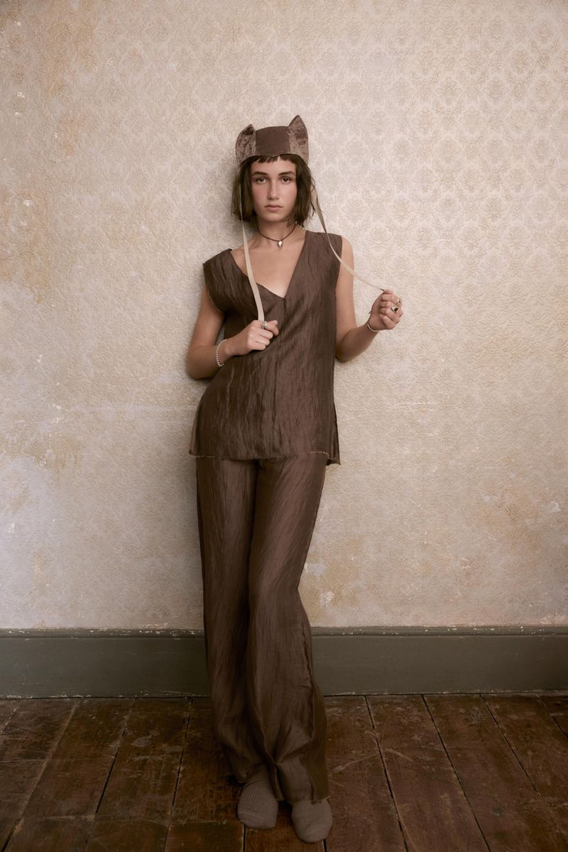 A young woman in a brown outfit and hat poses against a neutral patterned wallpaper. She is holding thin beige cords.